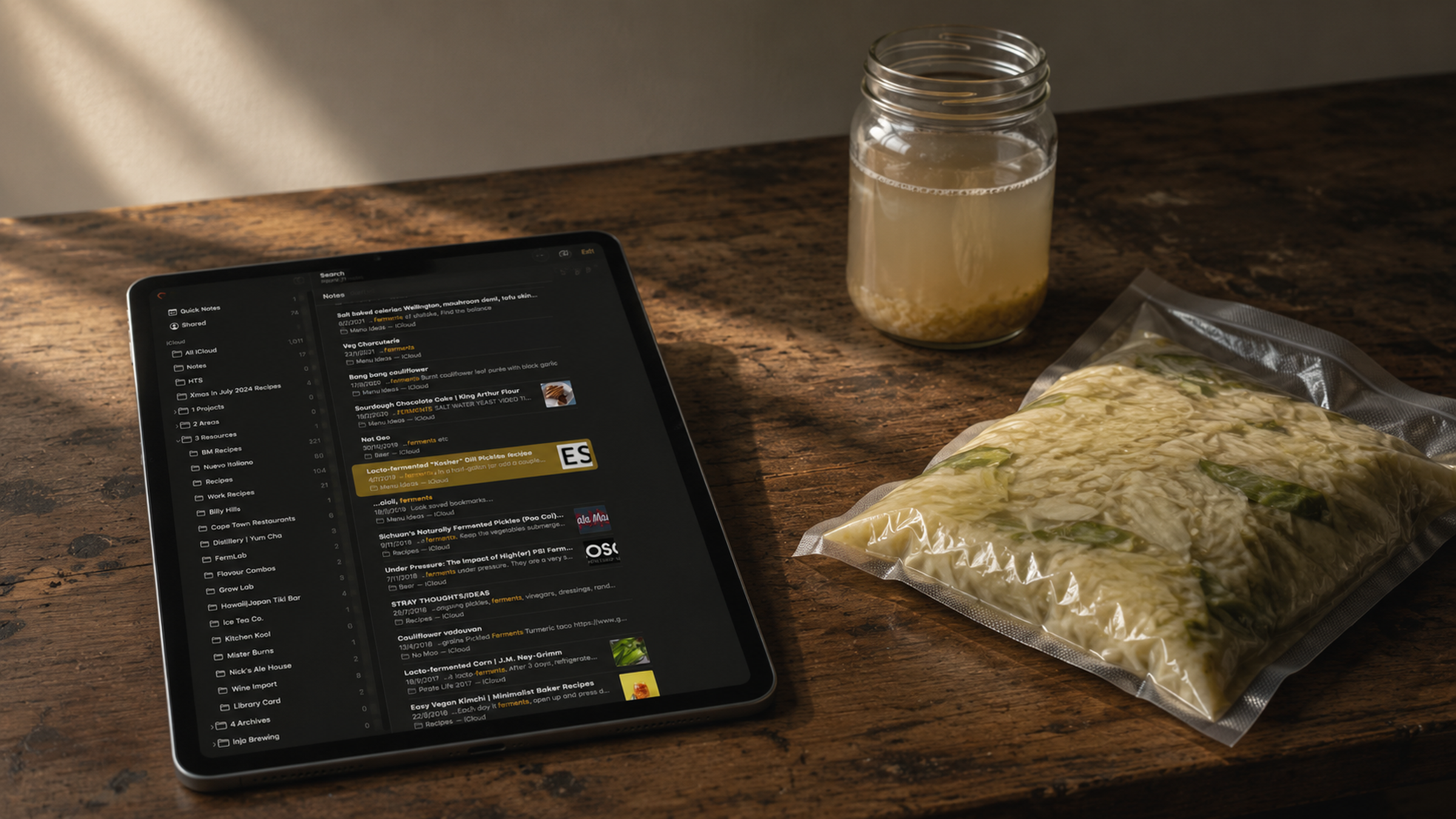 An iPad on a walnut bench showing a fermentation notes archive, beside a brine jar and a vacuum-sealed bag of cabbage in neutral-warm window light. The publication's writing surface.