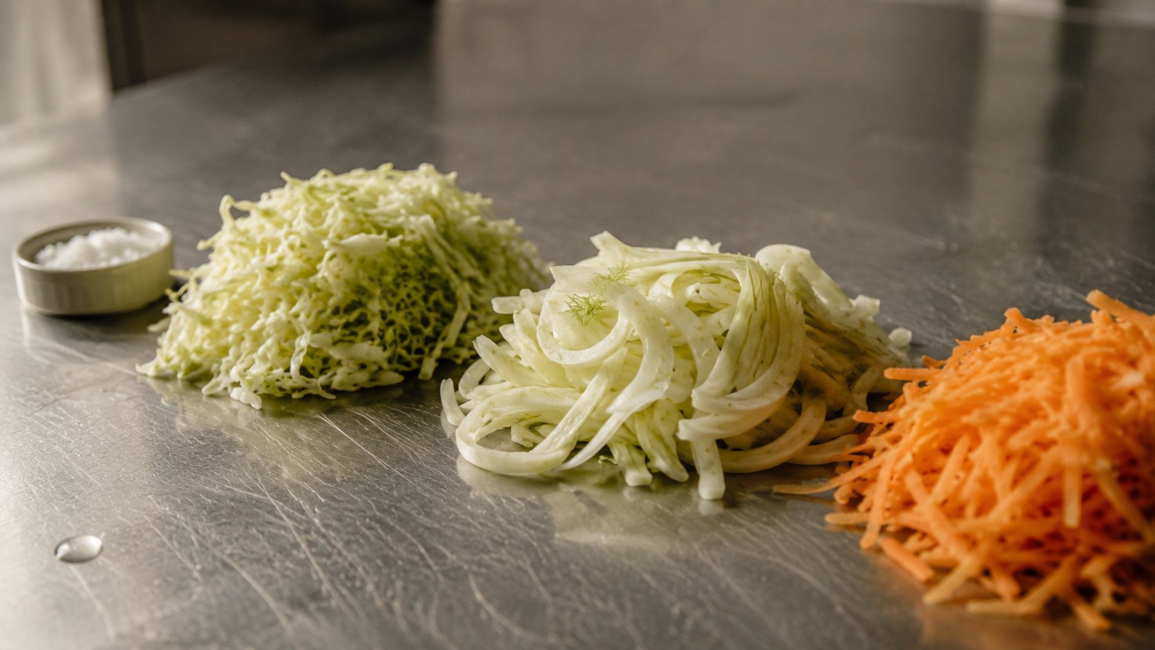 Shredded cabbage, sliced fennel and grated carrot with a ramekin of salt on a stainless bench