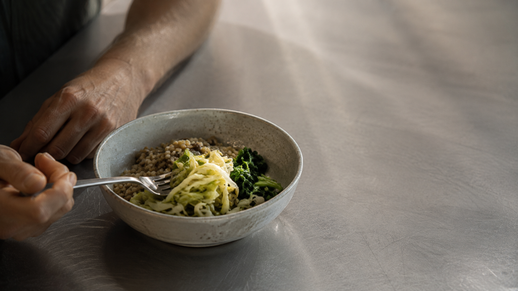 A bowl of grain, greens and fermented cabbage eaten unhurried on a steel bench, a relaxed hand resting beside it.