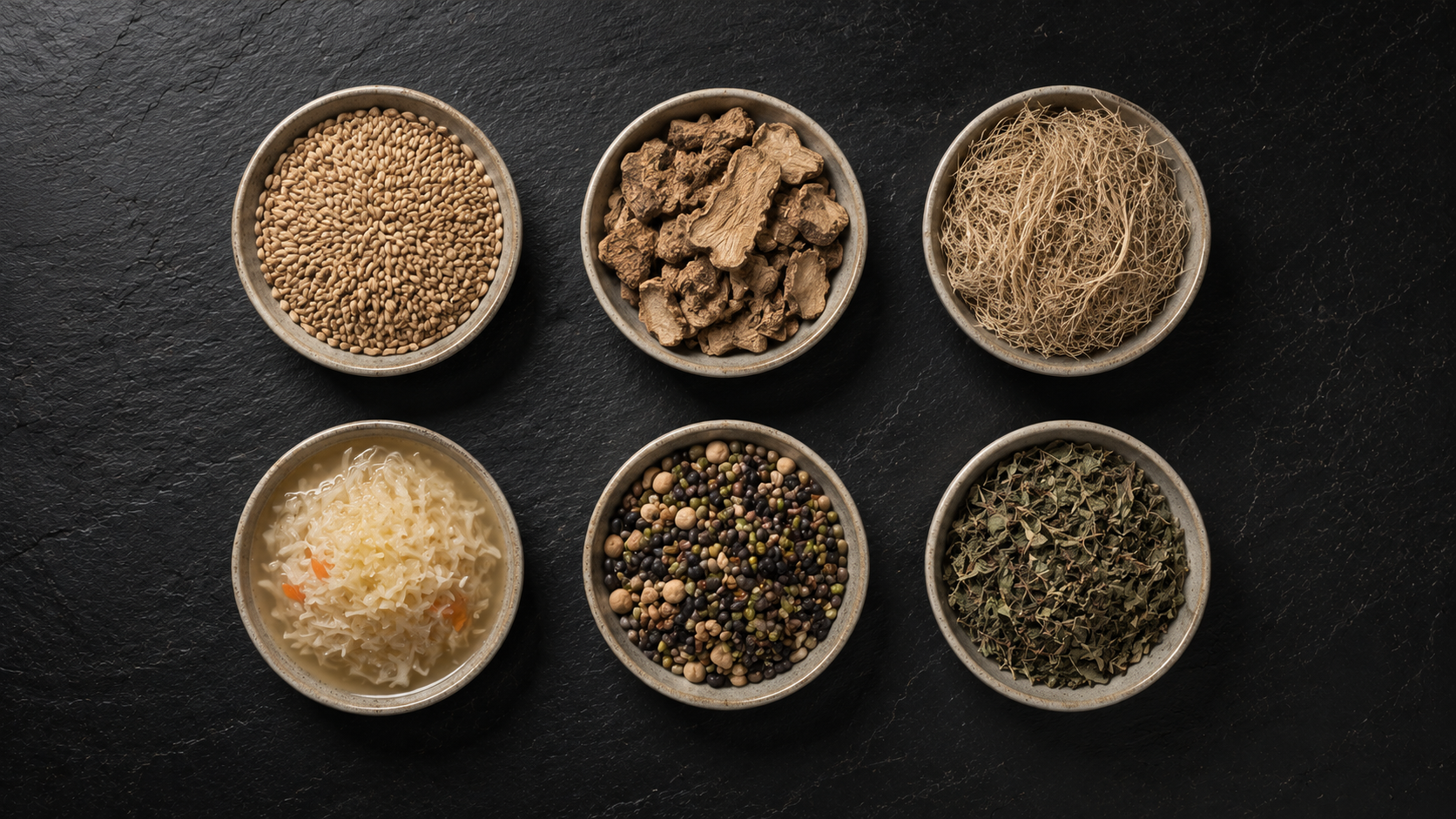 Six identical ceramic dishes on a dark slate surface, each containing a different natural specimen — grains, roots, fibres, fermented vegetables, seeds, dried herbs