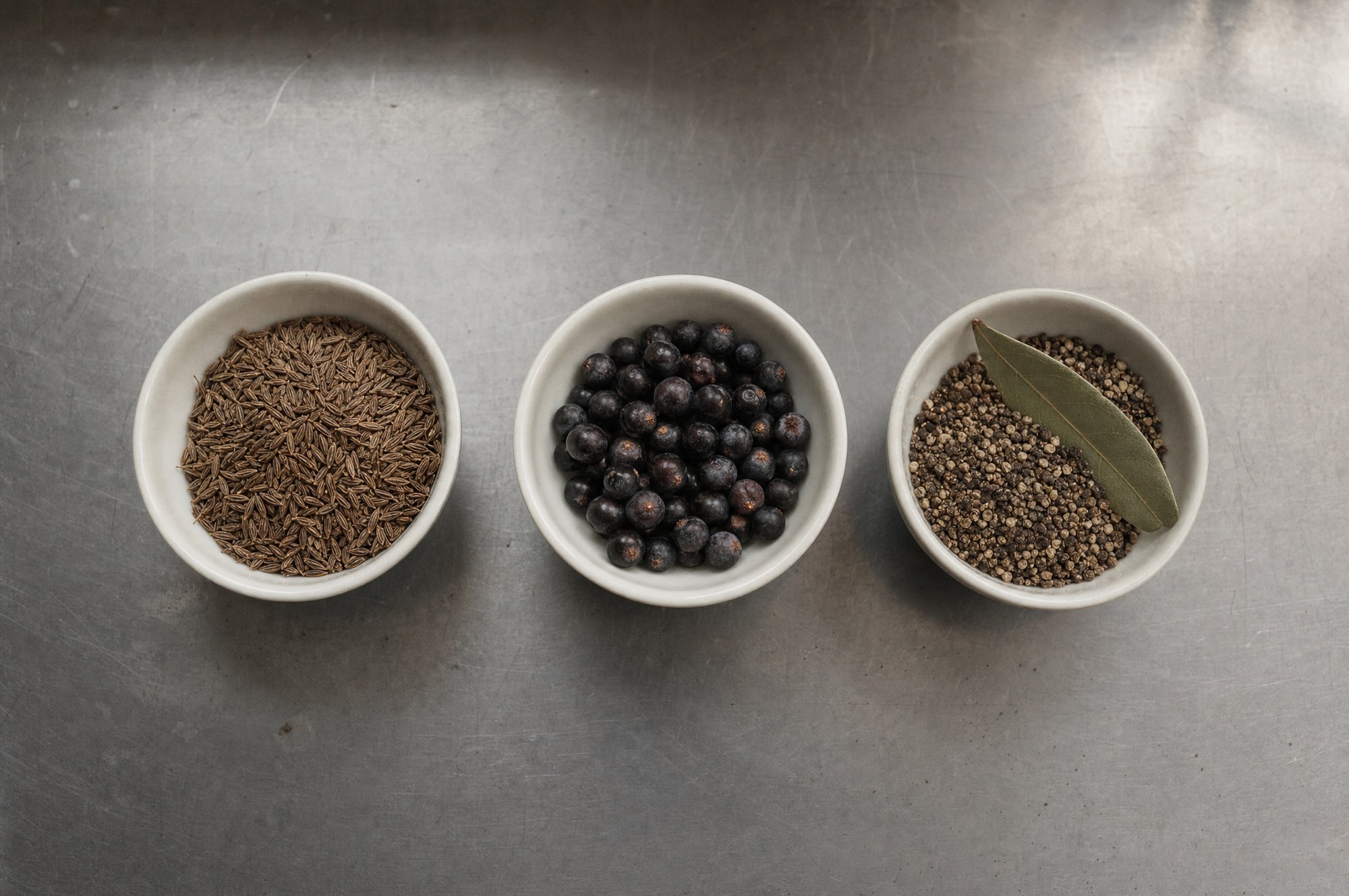 Three bowls of caraway, juniper and black pepper with a bay leaf on a steel bench