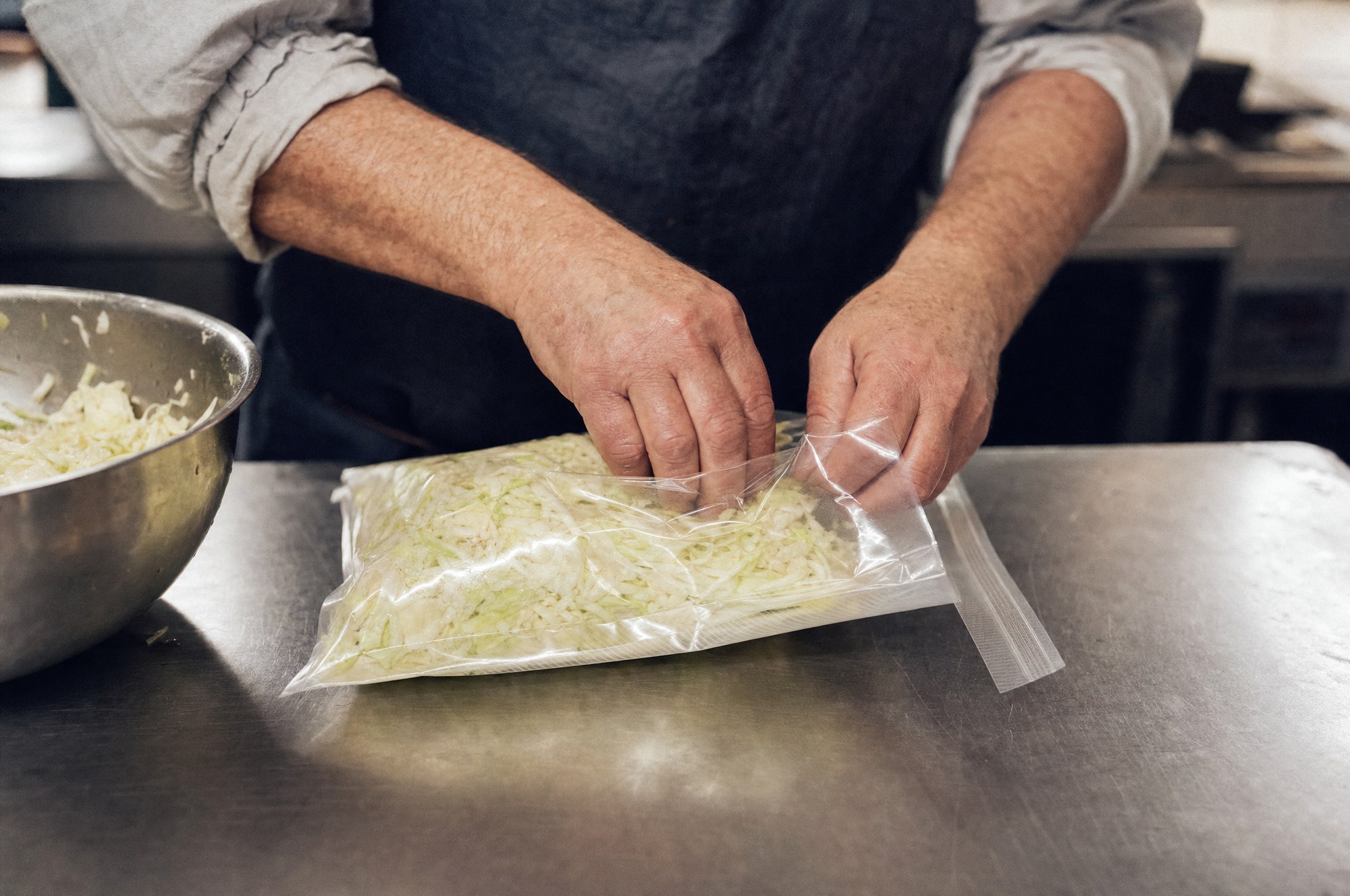 Hands packing salted cabbage into a vacuum bag on a steel bench