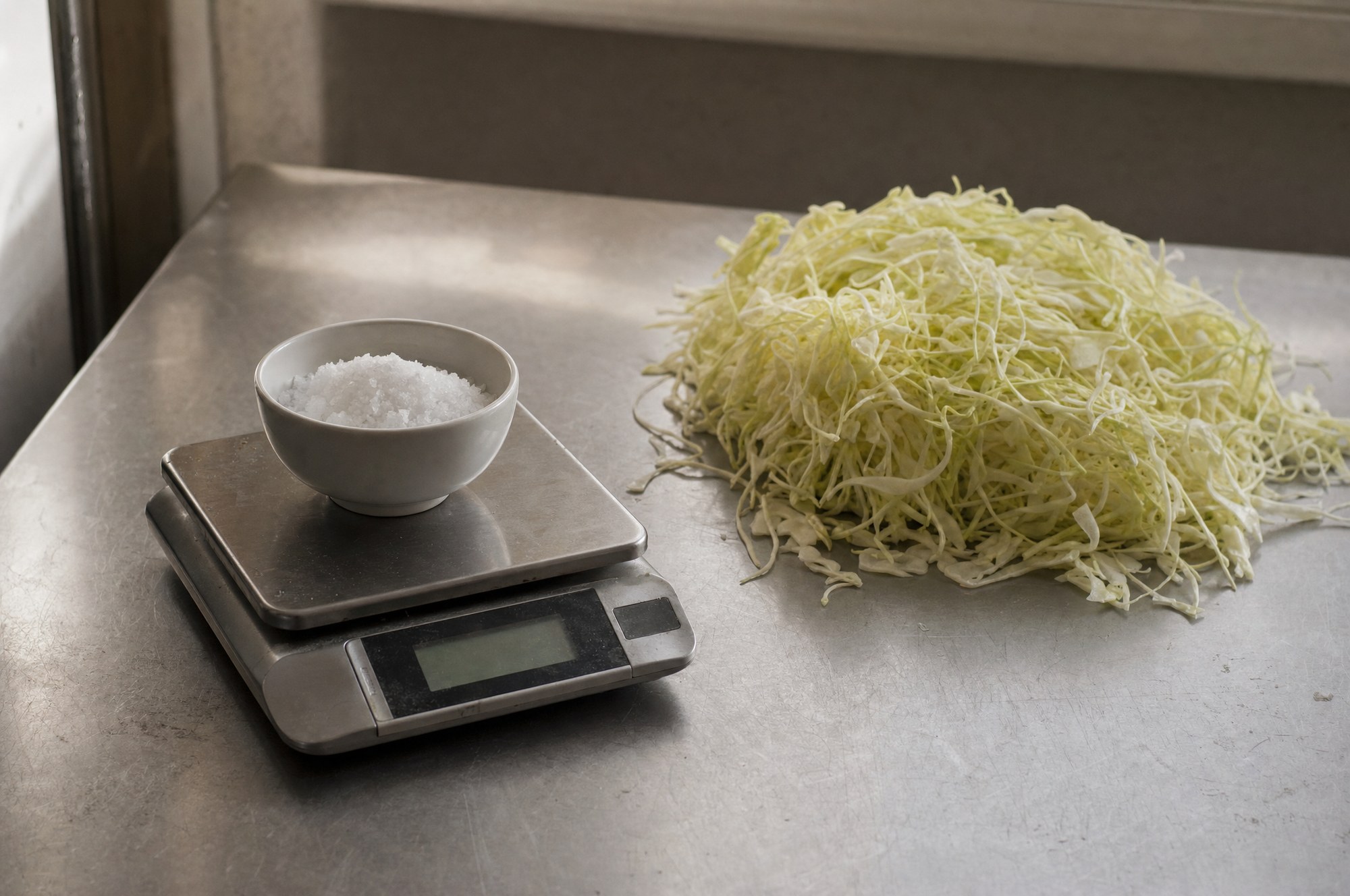 Bowl of salt on a digital scale beside shredded cabbage on a steel bench