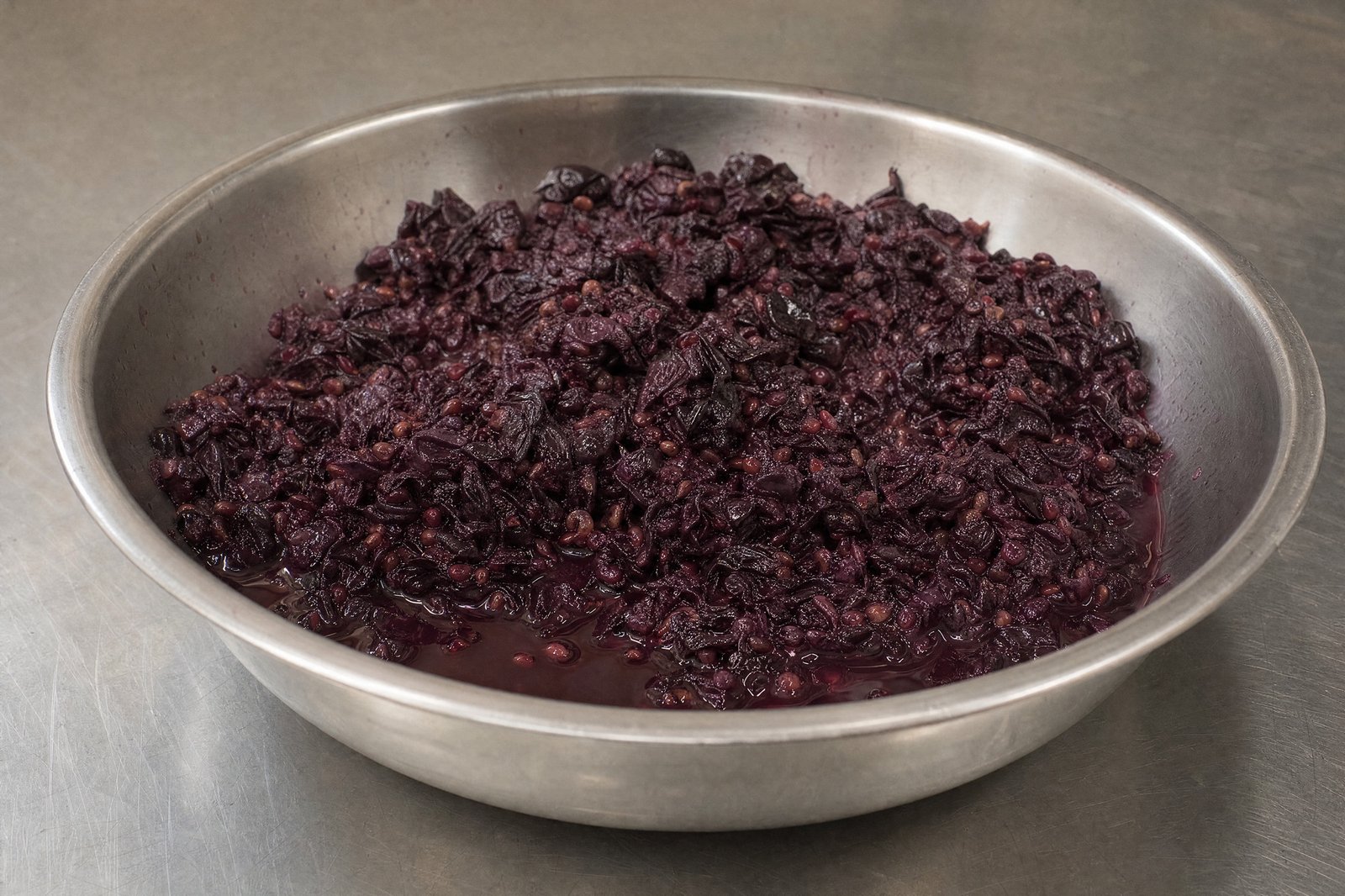 Pressed red wine grape skins and seeds in a steel bowl, dark pigment pooling beneath them